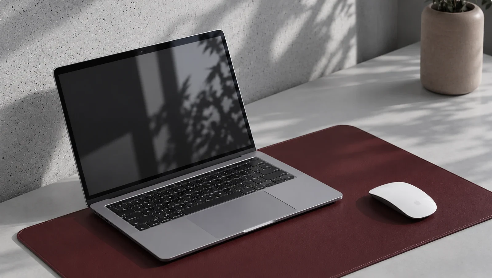 A silver laptop and white mouse on a burgundy desk mat in neutral daylight